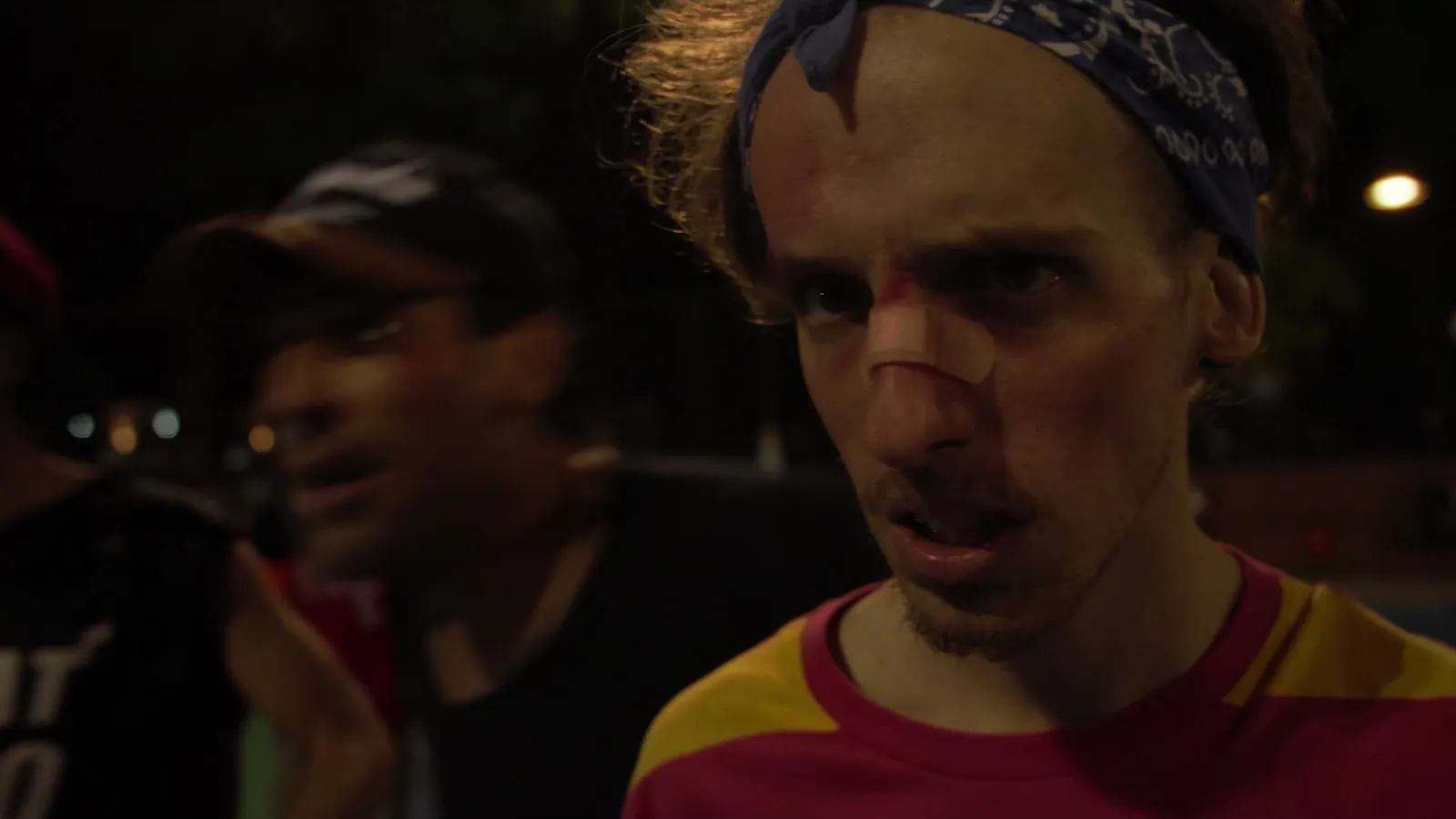 Close-up of Felipe with a blue headband and a bandaged nose; beside him, an out-of-focus rival