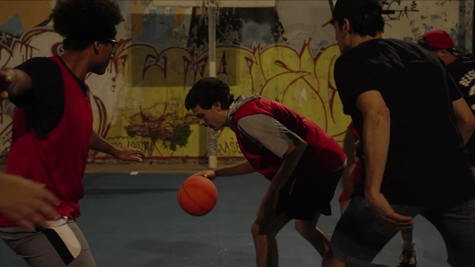 A player in a red jersey dribbles low between two rivals in black shirts, in front of a graffiti wall